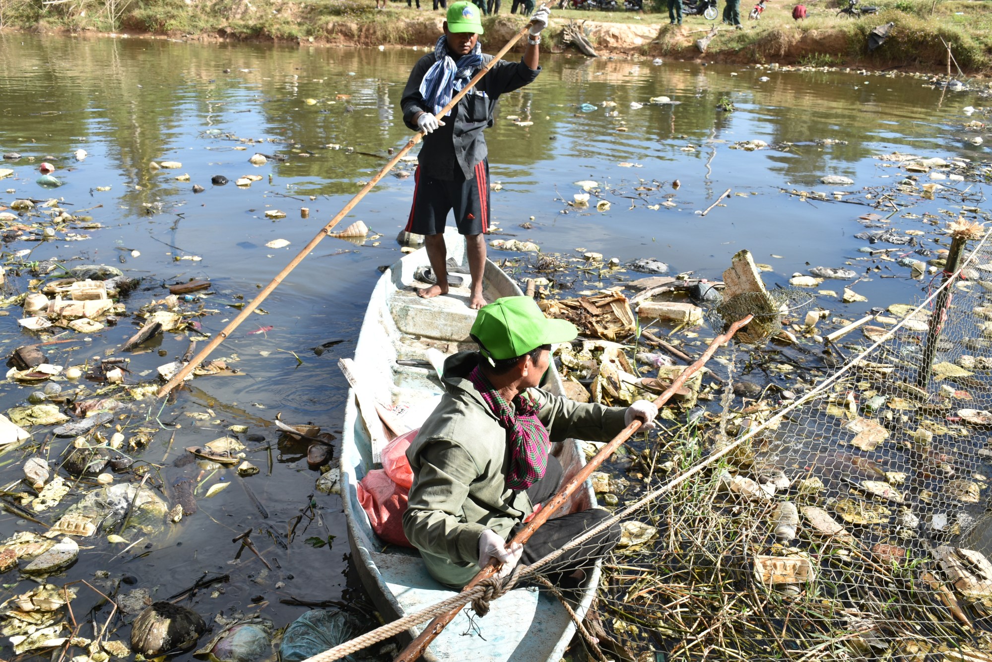Two people in a wooden boat surrounded by floating plastic