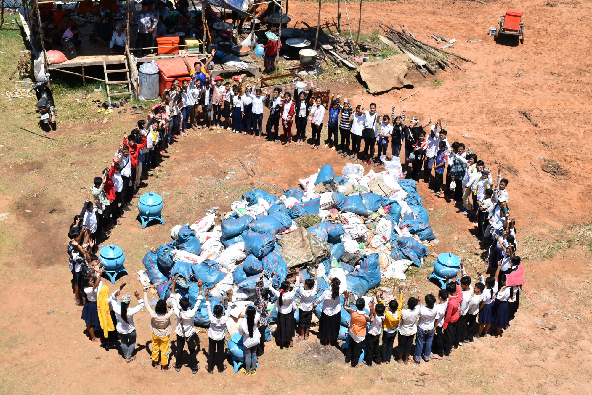 Seen from above, a large circle of students and villagers stands around bags of collected plastic